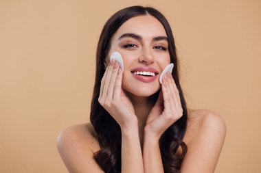 Güzel esmer, yarı çıplak bir kadın pamukla yüzünden makyaj malzemelerini çıkarıyor. Mutlu milenyum bayanı pamuk bezle deriyi temizliyor, bej rengi stüdyo arka planı, vesikalık resmi, kopyalama alanı