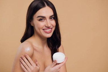 Gülen, yarı çıplak, gülümseyen ve bej arka planda izole edilmiş yüz kremi süren güzel bir kadının güzel portresini kapat.