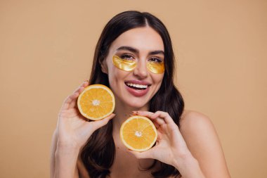 Güzel, saf, kibar bir kadının çıplak, izole bir şekilde duran görüntüsü. Gözlerinin altında altın bantlarla cildine dikkat et. Bej arka planda yarım portakal ile fotokopi alanı tut.