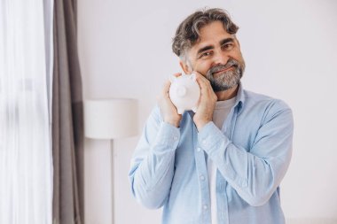 Sakallı yaşlı adam beyaz bir kumbara gösteriyor. Tasarruf temaları, emeklilik ve fotokopi alanı ile finansal planlama öneriyor.