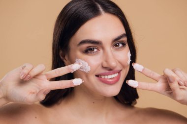 Kadın güzel, mutlu yüz, krem sürerek ve cildini temizlerken parmakları, tırnağı ve saçı, güzel saçı, vücudu ve bej arka planda güzellik konsepti.