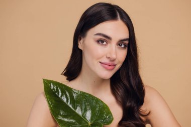 Temiz, parlak yüzlü, bej arka planda yeşil yapraklı genç bir kadın. Spa, yüz bakımı, güzellik kozmetolojisi. Doğal kozmetik konsepti.