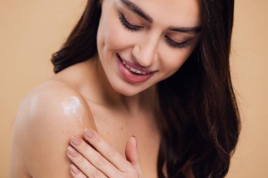 Gülen, yarı çıplak, gülümseyen ve bej arka planda izole edilmiş yüz kremi süren güzel bir kadının güzel portresini kapat.