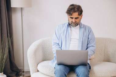 Oturma odasında dizüstü bilgisayar kullanarak gülümseyen olgun bir adam. Mutlu kendinden emin yetişkin erkek evde dinleniyor bilgisayar üzerinde çalışıyor, klavyede daktilo yazıyor, boşluğu kopyalıyor