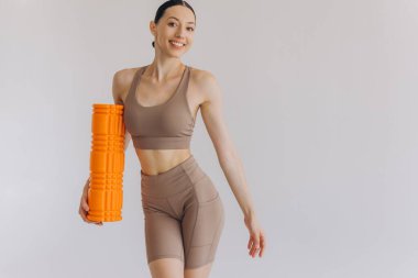 Bej rengi spor giysili zayıf bir kadının portresi spor salonu için turuncu köpük silindirli güvenli bir şekilde duruyor, fotokopi alanı ile beyaz üzerinde poz veriyor, sağlık ve refahı destekliyor..