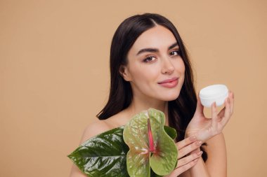 Yeşil çiçeğin yanındaki çekici bir kadının portresi sorun için yüz kremi sürüyor. Bej arka planda merhem var. Refah konsepti