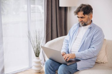 Oturma odasında dizüstü bilgisayar kullanarak gülümseyen olgun bir adam. Mutlu kendinden emin yetişkin erkek evde dinleniyor bilgisayar üzerinde çalışıyor, klavyede daktilo yazıyor, boşluğu kopyalıyor