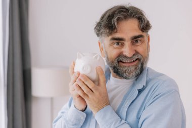 Sakallı yaşlı adam beyaz bir kumbara gösteriyor. Tasarruf temaları, emeklilik ve fotokopi alanı ile finansal planlama öneriyor.