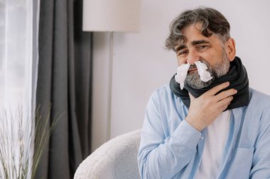 Gri saçlı hasta bir adam boğazını tutuyor ve burnunda da ilaç reklamı için fotokopi yeri olan bir burun peçetesi var.