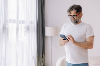 Beyaz tişörtlü, elinde cep telefonu olan ve kameraya gülümseyen yaşlı gri saçlı bir adamın portresi..