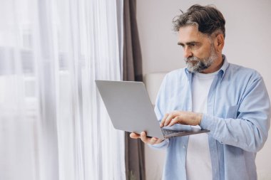 Oturma odasında dizüstü bilgisayar kullanarak gülümseyen olgun bir adam. Mutlu kendinden emin yetişkin erkek evde dinleniyor bilgisayar üzerinde çalışıyor, klavyede daktilo yazıyor, boşluğu kopyalıyor