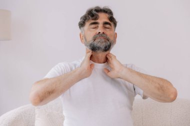 Gri saçlı adamın boğazı ağrıyormuş. Yaşlı bir erkek koltukta oturup elini boğaz iltihabından tutmuş ve boğazı ağrıyormuş..
