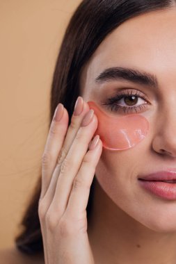 Göz bantları. Güzellik konsepti. Güzel bir model gözlerine pembe hidro jel sürerek bej arka planda gülümsüyor.