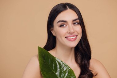 Temiz, parlak yüzlü, bej arka planda yeşil yapraklı genç bir kadın. Spa, yüz bakımı, güzellik kozmetolojisi. Doğal kozmetik konsepti.