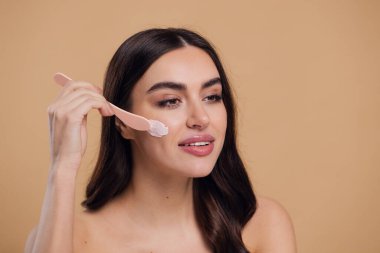 Yarı çıplak kadın, güzellik için yüz ve fırça kremi, kuaför ve cilt bakımı bej arka planda. Mutlu model, yüz losyonu ve dermatoloji, kolajen ve mutluluğun gülümsemesi için makyaj malzemesi.