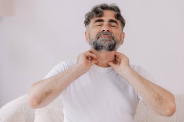 Gri saçlı adamın boğazı ağrıyormuş. Yaşlı bir erkek koltukta oturup elini boğaz iltihabından tutmuş ve boğazı ağrıyormuş..