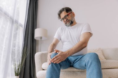 Ev, diz ağrısı ve koltukta eklem iltihabı, kemik erimesi ve fibromiyalji olan yaşlı bir adam. Rahatlama, kas gerilimi ve kaza ya da emeklilikte yaralanma için eklem masajı yapan yaşlı erkek.