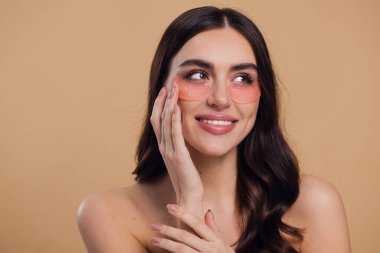 Göz bantları. Güzellik konsepti. Güzel bir model gözlerine pembe hidro jel sürerek bej arka planda gülümsüyor.