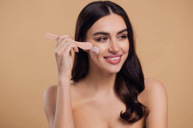 Yarı çıplak kadın, güzellik için yüz ve fırça kremi, kuaför ve cilt bakımı bej arka planda. Mutlu model, yüz losyonu ve dermatoloji, kolajen ve mutluluğun gülümsemesi için makyaj malzemesi.