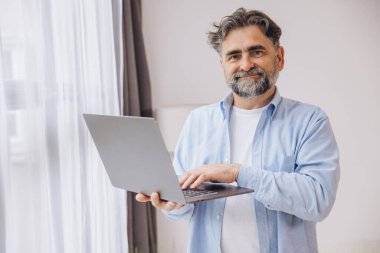Oturma odasında dizüstü bilgisayar kullanarak gülümseyen olgun bir adam. Mutlu kendinden emin yetişkin erkek evde dinleniyor bilgisayar üzerinde çalışıyor, klavyede daktilo yazıyor, boşluğu kopyalıyor