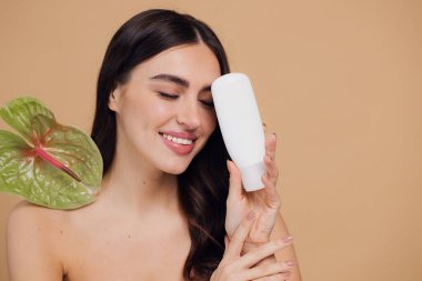 Yeşil çiçeğin yanındaki çekici bir kadının portresi sorun için yüz kremi sürüyor. Bej arka planda merhem var. Refah konsepti