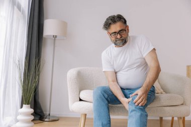 Ev, diz ağrısı ve koltukta eklem iltihabı, kemik erimesi ve fibromiyalji olan yaşlı bir adam. Rahatlama, kas gerilimi ve kaza ya da emeklilikte yaralanma için eklem masajı yapan yaşlı erkek.