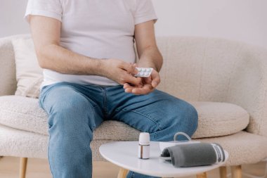 Close-up of fat elderly man sitting sofa at living room measures blood pressure with device. Senior male holding pills and drops drugs. Health and self-care concept, copy space clipart