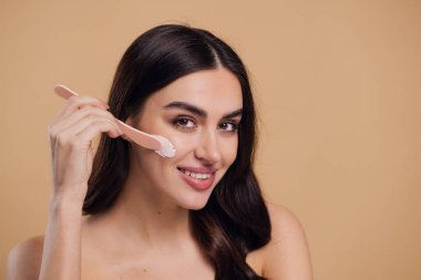 Yarı çıplak kadın, güzellik için yüz ve fırça kremi, kuaför ve cilt bakımı bej arka planda. Mutlu model, yüz losyonu ve dermatoloji, kolajen ve mutluluğun gülümsemesi için makyaj malzemesi.