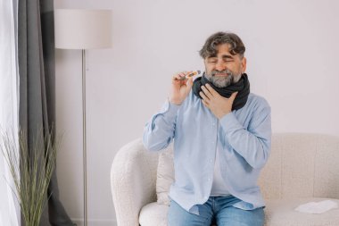 Yüksek ateşi olan yaşlı bir adam, evdeyken termometresini dikkatlice kontrol ediyor. Etrafı çeşitli ilaçlarla çevrili bir battaniyeye sarılmış ve kendini pek iyi hissetmiyor ve endişeli görünüyor..