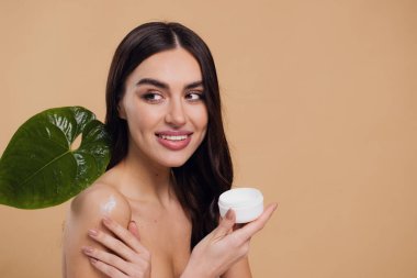 Gülen, yarı çıplak, gülümseyen ve bej arka planda izole edilmiş yüz kremi süren güzel bir kadının güzel portresini kapat.