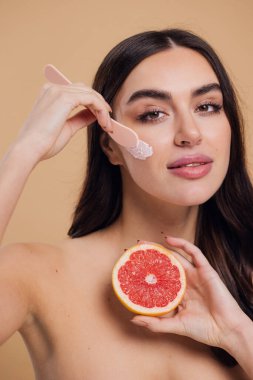 Yarı çıplak kadın, güzellik için yüz ve fırça kremi, kuaför ve cilt bakımı bej arka planda. Mutlu model gülümsüyor ve elinde greyfurt, yüz losyonu ve dermatoloji için makyaj malzemesi tutuyor.