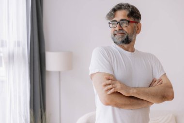 Mutlu, kendinden emin, gözlüklü, kolları bağlı yaşlı bir adam. Yaşlı olgun gülümseyen 60 yaşındaki yakışıklı adam modern evdeki oturma odasında poz veriyor. Portre.