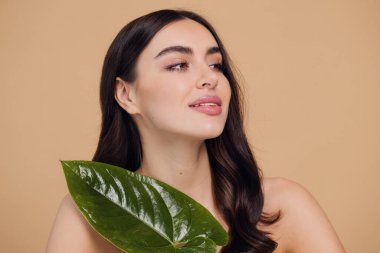Temiz, parlak yüzlü, bej arka planda yeşil yapraklı genç bir kadın. Spa, yüz bakımı, güzellik kozmetolojisi. Doğal kozmetik konsepti.