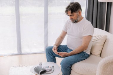 Oturma odasındaki koltukta oturan yaşlı adam kan basıncını cihazla ölçüyor. Yaşlı erkek hapları tutuyor ve uyuşturucu bırakıyor. Sağlık ve öz bakım kavramı, kopyalama alanı