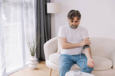 Evdeki kanepede oturmuş, tansiyonunu ölçen, gri saçlı, son sınıf öğrencisi. Modern tonometre kullanan emekli, hipertansiyon deneyimi yaşayan, kopyalama alanı