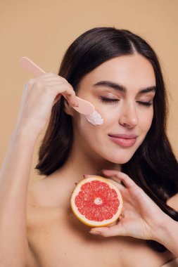 Yarı çıplak kadın, güzellik için yüz ve fırça kremi, kuaför ve cilt bakımı bej arka planda. Mutlu model gülümsüyor ve elinde greyfurt, yüz losyonu ve dermatoloji için makyaj malzemesi tutuyor.
