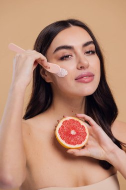 Yarı çıplak kadın, güzellik için yüz ve fırça kremi, kuaför ve cilt bakımı bej arka planda. Mutlu model gülümsüyor ve elinde greyfurt, yüz losyonu ve dermatoloji için makyaj malzemesi tutuyor.