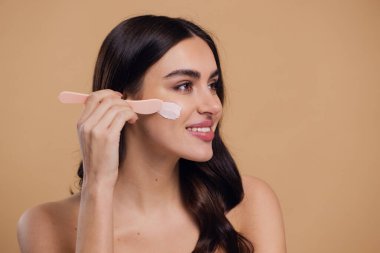 Yarı çıplak kadın, güzellik için yüz ve fırça kremi, kuaför ve cilt bakımı bej arka planda. Mutlu model, yüz losyonu ve dermatoloji, kolajen ve mutluluğun gülümsemesi için makyaj malzemesi.