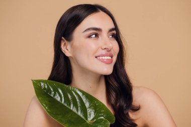 Temiz, parlak yüzlü, bej arka planda yeşil yapraklı genç bir kadın. Spa, yüz bakımı, güzellik kozmetolojisi. Doğal kozmetik konsepti.