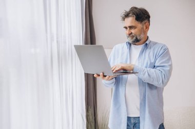 Oturma odasında dizüstü bilgisayar kullanarak gülümseyen olgun bir adam. Mutlu kendinden emin yetişkin erkek evde dinleniyor bilgisayar üzerinde çalışıyor, klavyede daktilo yazıyor, boşluğu kopyalıyor