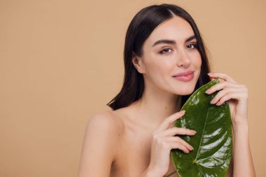Temiz, parlak yüzlü, bej arka planda yeşil yapraklı genç bir kadın. Spa, yüz bakımı, güzellik kozmetolojisi. Doğal kozmetik konsepti.