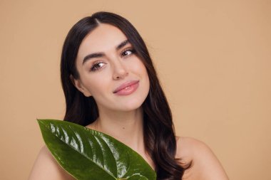 Temiz, parlak yüzlü, bej arka planda yeşil yapraklı genç bir kadın. Spa, yüz bakımı, güzellik kozmetolojisi. Doğal kozmetik konsepti.
