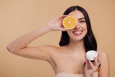 Kadın, doğal cilt bakımı için nemlendirici krem ve turuncu ve bej renkli stüdyo arka planına karşı C vitamini. Sağlıklı organik ürünler için meyve ve krem ya da losyon tutan mutlu kadın portresi