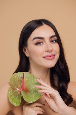 Bej renkli stüdyo arka planında izole edilmiş çiçekle poz veren çekici genç bir kadının güzel portresi.