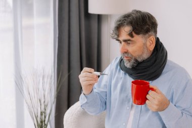 Yüksek ateşi olan yaşlı bir adam, termometresini dikkatlice kontrol ediyor ve evdeyken bir fincan çay içiyor. Etrafı çeşitli ilaçlarla çevrili bir battaniyeye sarılmış ve kendini pek iyi hissetmiyor gibi görünüyor..