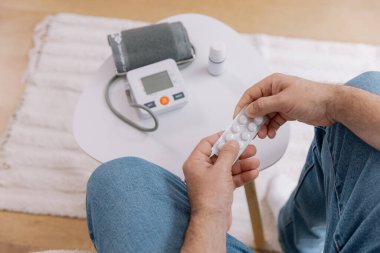 Oturma odasındaki koltukta oturan yaşlı adamın yakın çekimi kan basıncını cihazla ölçüyor. Yaşlı erkek hapları tutuyor ve uyuşturucu bırakıyor. Sağlık ve öz bakım kavramı, kopyalama alanı