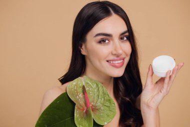 Yeşil çiçeğin yanındaki çekici bir kadının portresi sorun için yüz kremi sürüyor. Bej arka planda merhem var. Refah konsepti