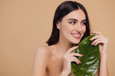 Temiz, parlak yüzlü, bej arka planda yeşil yapraklı genç bir kadın. Spa, yüz bakımı, güzellik kozmetolojisi. Doğal kozmetik konsepti.