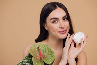 Yeşil çiçeğin yanındaki çekici bir kadının portresi sorun için yüz kremi sürüyor. Bej arka planda merhem var. Refah konsepti