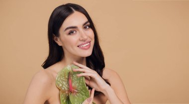 Bej renkli stüdyo arka planında izole edilmiş çiçekle poz veren çekici genç bir kadının güzel portresi.